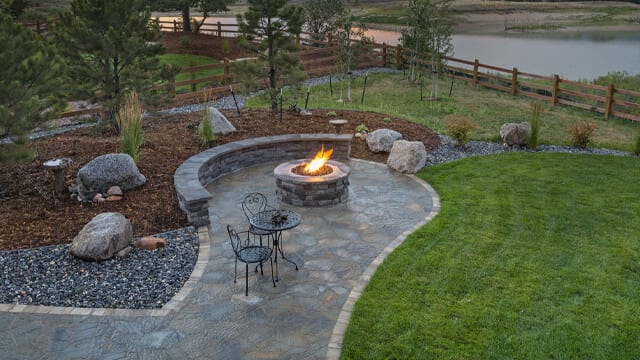 An outdoor firepit surrounded by a patio area and rock and pine mulch. Landscaping boulder add an accent to the landscaping.